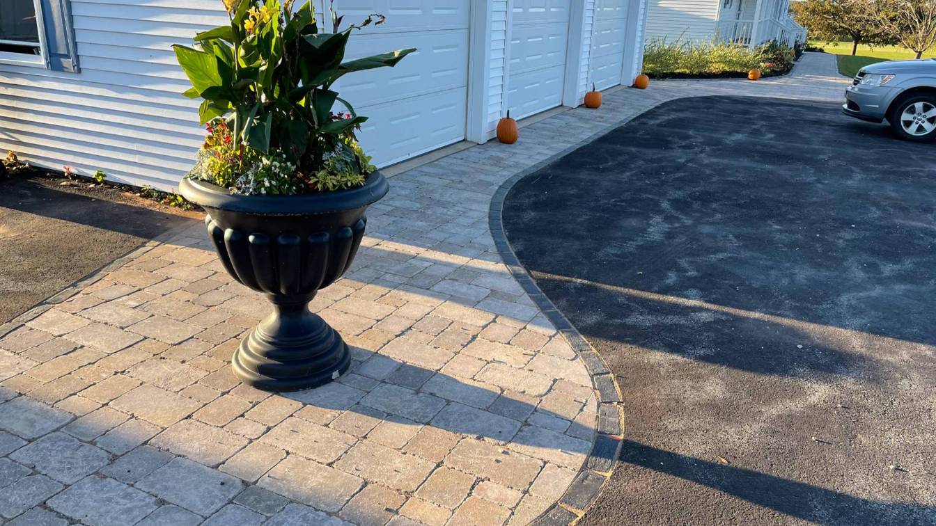 Paver walkway with curved edges, black decorative planter filled with flowers, and garage doors in a residential setting, highlighting hardscaping services by McIsaac Lawncare in Prince Edward Island.