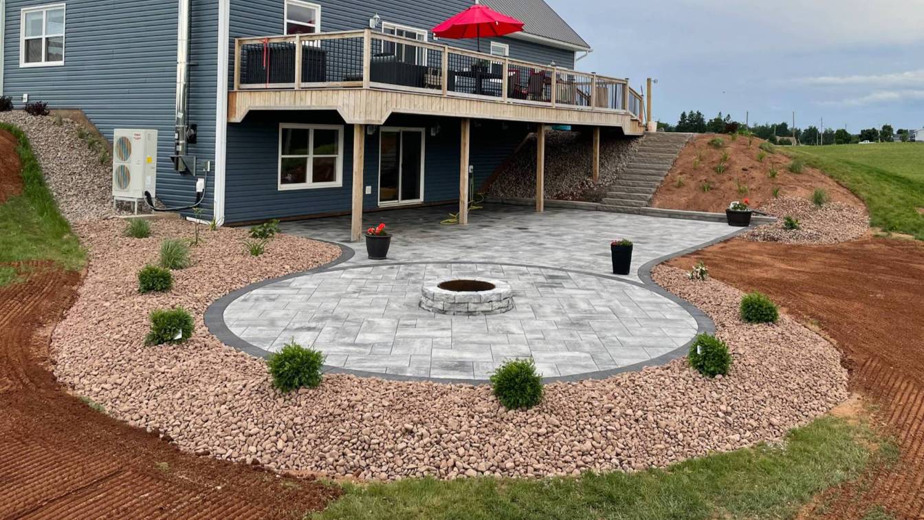 Paver fire pit surrounded by a circular stone patio, decorative plants, and gravel landscaping, enhancing outdoor living space for warmth and ambiance.