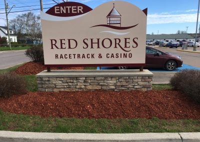 Signage for Red Shores Racetrack & Casino, featuring the words "ENTER" and "RED SHORES RACETRACK & CASINO," with landscaped flower beds and mulch in the foreground, illustrating a welcoming entrance to the facility.