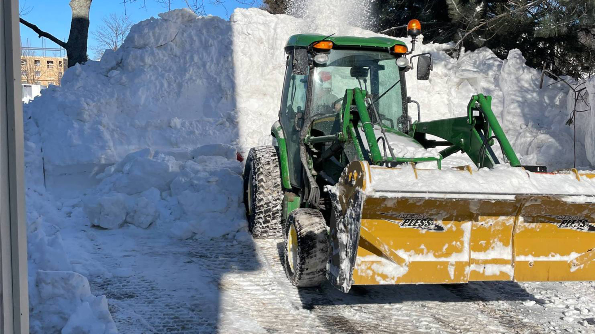 Snow removal equipment clearing a pathway, featuring a green tractor with a yellow snow plow, surrounded by large snow piles against a clear blue sky, illustrating McIsaac Lawncare's snow removal services.