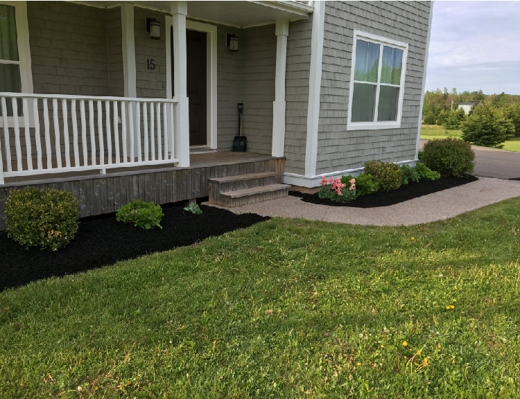 Front porch of a home with landscaped yard featuring green grass, flower beds, and black mulch, illustrating proper yard preparation for a healthy lawn.