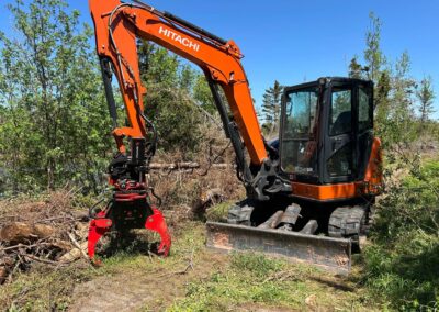 Hitachi excavator clearing overgrown lot with grapple attachment, showcasing landscaping transformation capabilities.