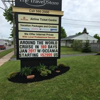 Sign for The Travel Store displaying vacation packages and cruise offers, featuring a well-maintained lawn and flowerbed, emphasizing local professional services.