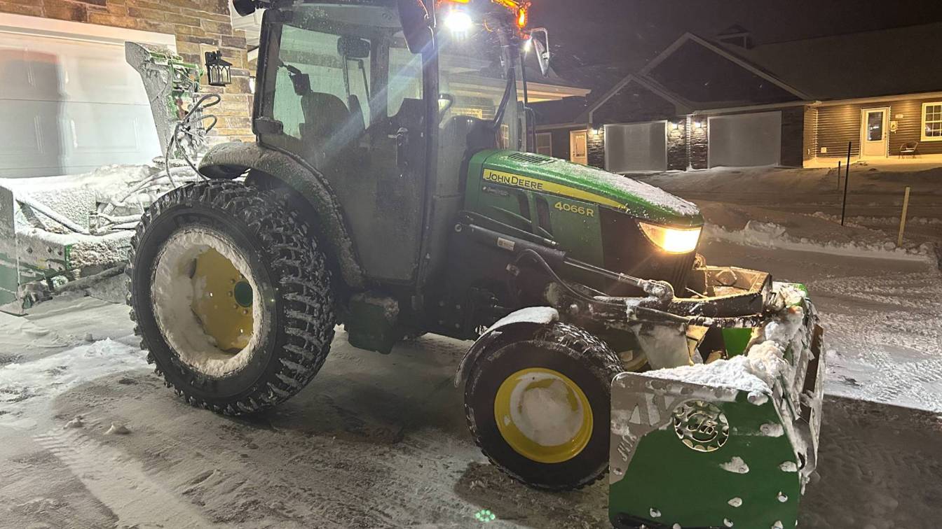 John Deere tractor equipped with a snow plow, covered in snow, parked in a residential area at night, highlighting snow removal services for driveways and parking lots.