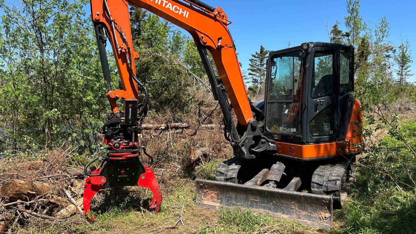 Excavator with a grapple attachment in a wooded area, used for tree and brush removal, showcasing McIsaac Lawncare's land clearing services.