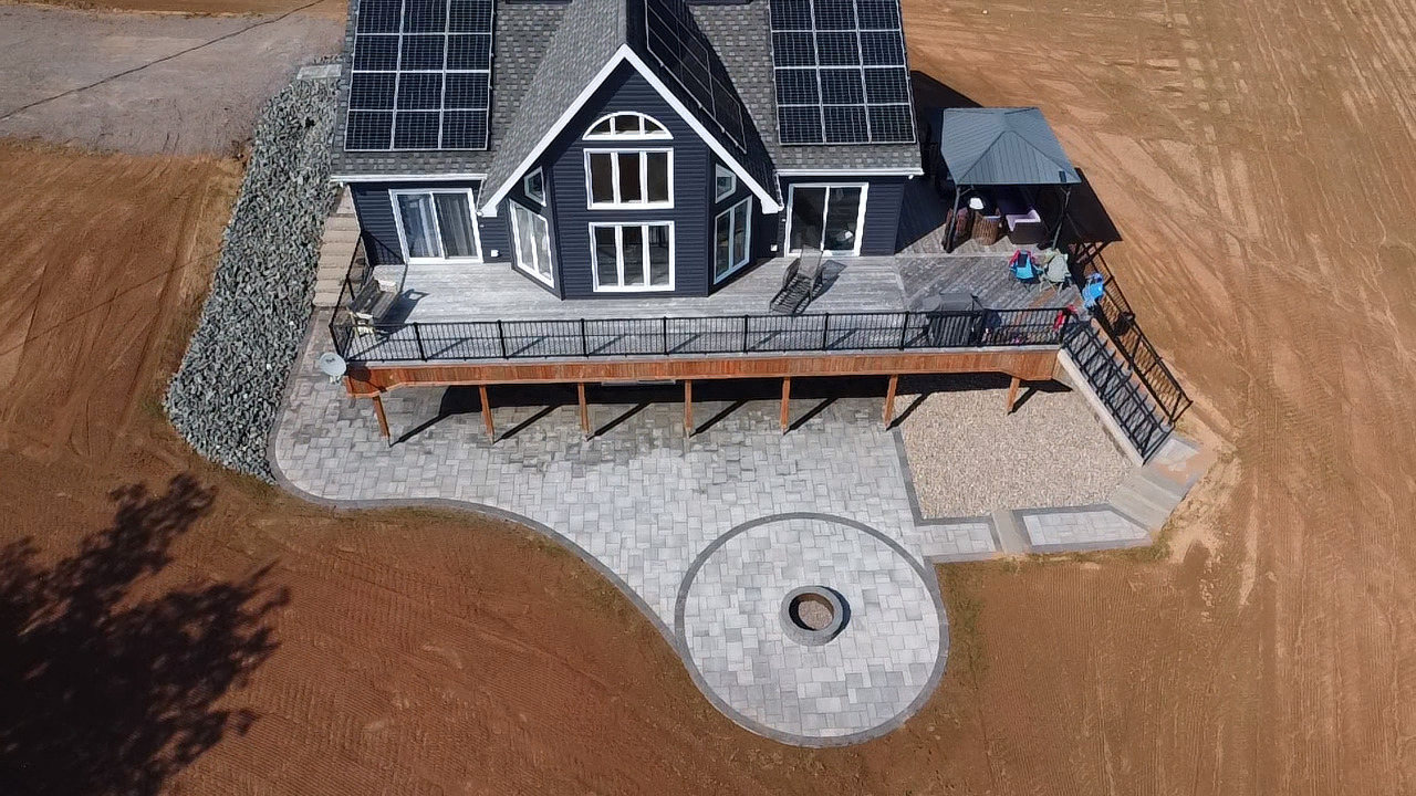 Aerial view of a modern home with solar panels, featuring a spacious paver patio with circular fire pit, surrounded by a landscaped area, showcasing custom hardscaping design by McIsaac Lawncare.