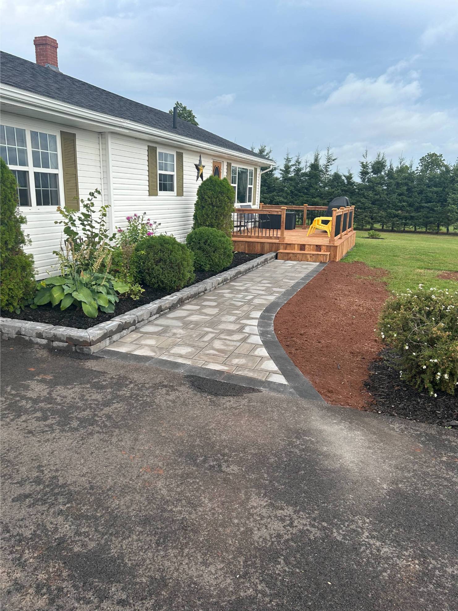 Paved walkway leading to a wooden deck, surrounded by landscaped greenery and flower beds, enhancing outdoor space aesthetics for hardscaping services.