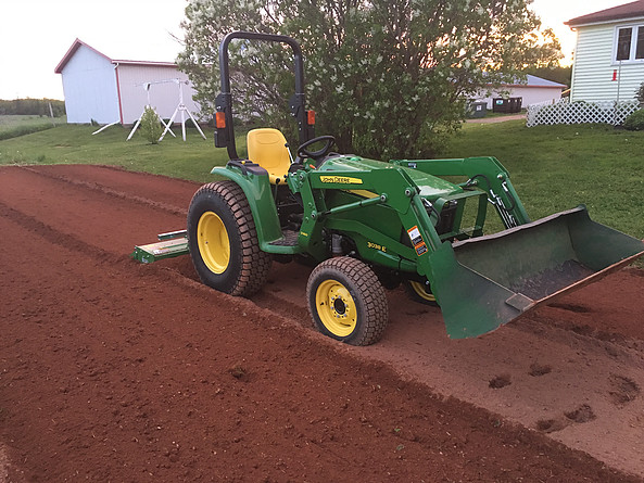 Green tractor preparing soil for traditional grass seeding in a residential yard, with nearby trees and houses visible.