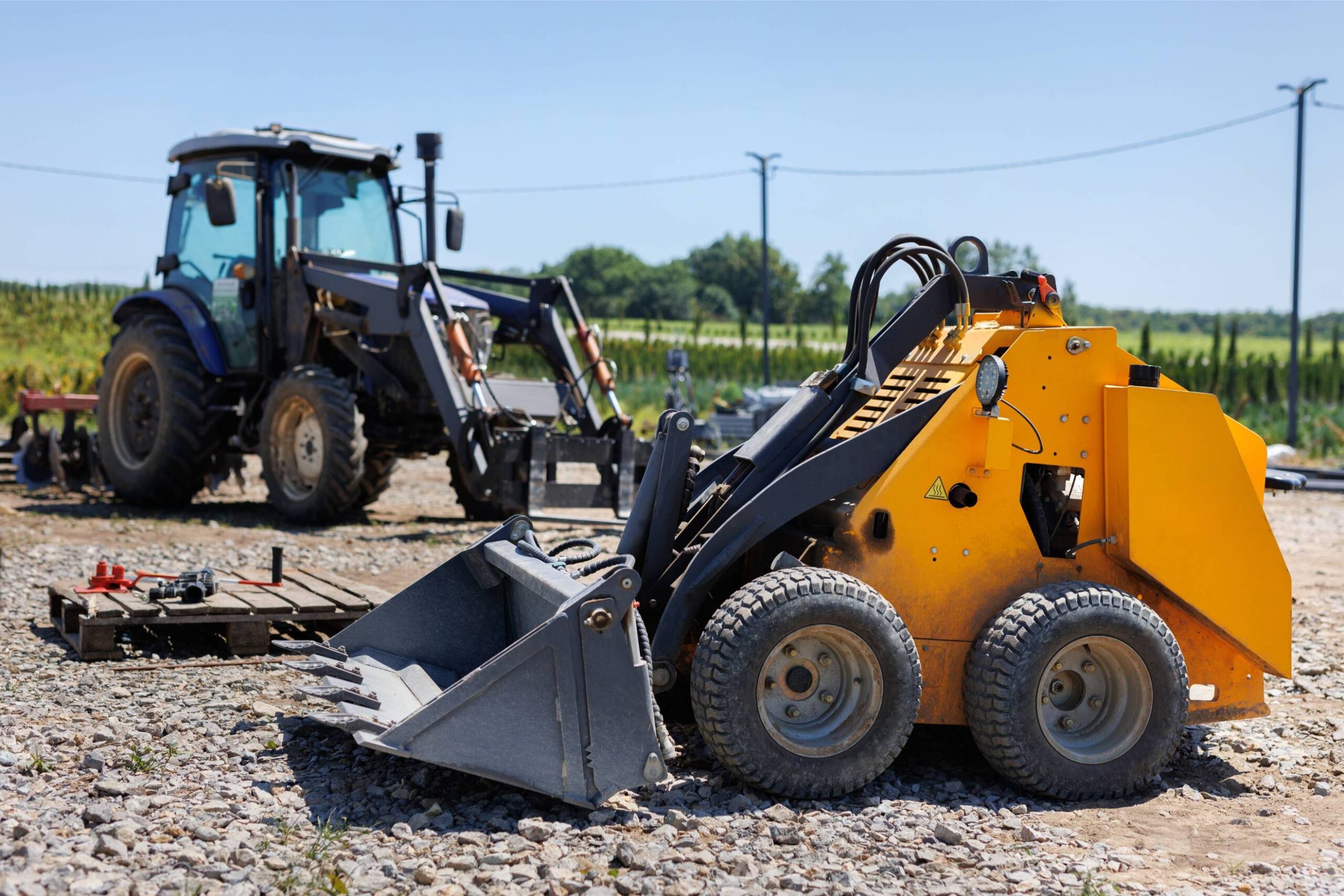 Small yellow skid steer loader with bucket, positioned on gravel ground, with a blue tractor and green fields in the background, illustrating excavation and trenching services.