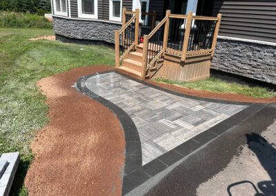 Paved walkway leading to a house entrance, showcasing recent landscaping work by McIsaac Lawncare, featuring gray stone tiles, bordered by black edging and brown soil.