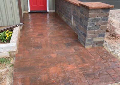 Paved walkway with textured red stone and gray stone wall, leading to a red door, showcasing McIsaac Lawncare's landscaping craftsmanship.