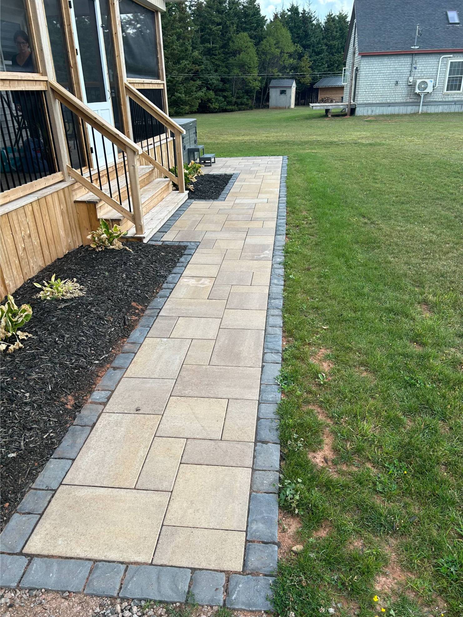 Paved walkway featuring interlocking stone design, bordered by dark mulch and plants, leading to a wooden deck, showcasing professional hardscaping services by McIsaac Lawncare.