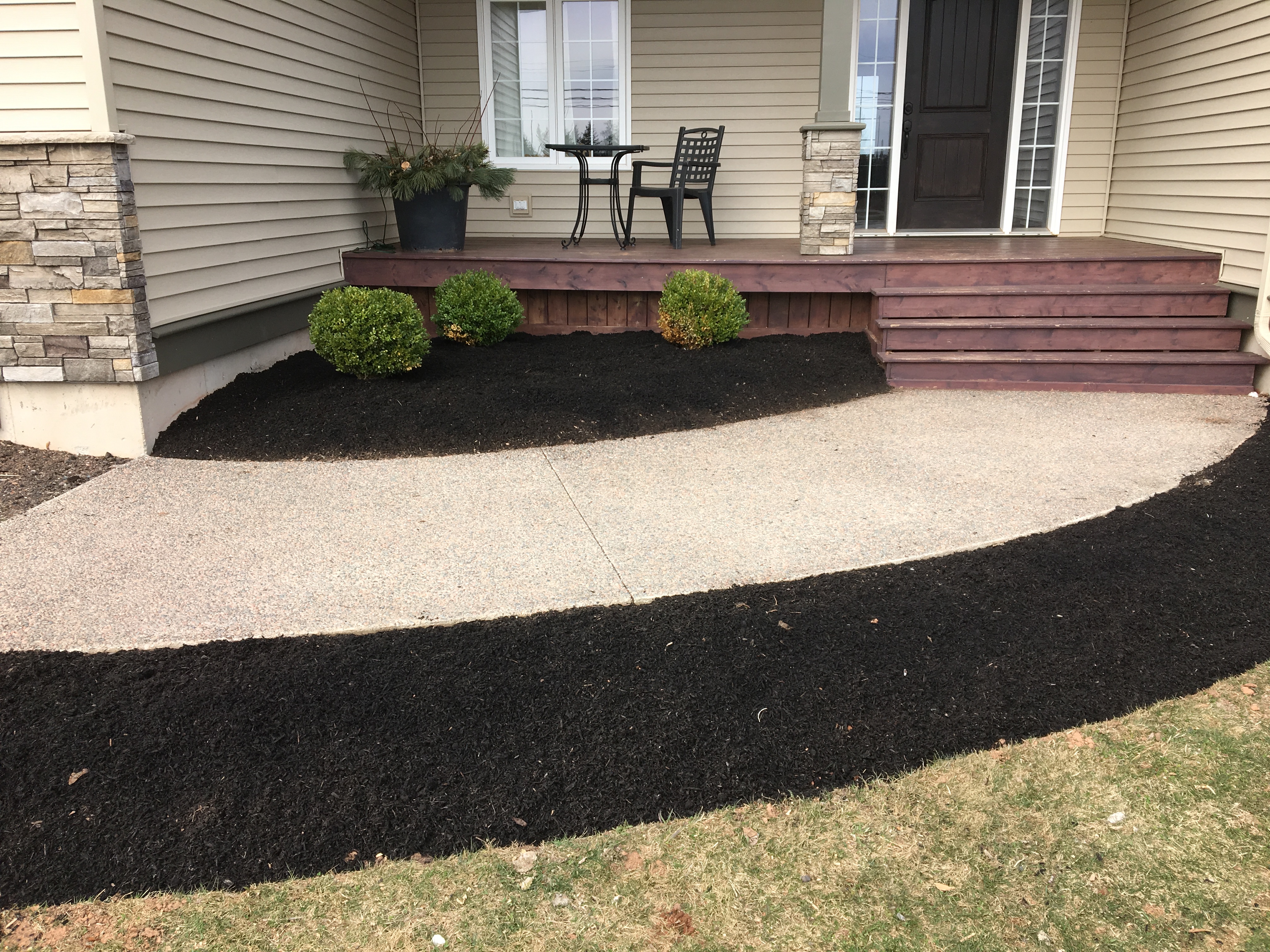 Custom patio area with curved concrete pathway, bordered by black mulch and small green shrubs, adjacent to wooden steps and a seating area, enhancing outdoor living space.