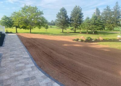 Landscaped backyard transformation featuring freshly prepared soil, bordered by paver stones, surrounded by greenery and trees, showcasing McIsaac Lawncare's quality craftsmanship.