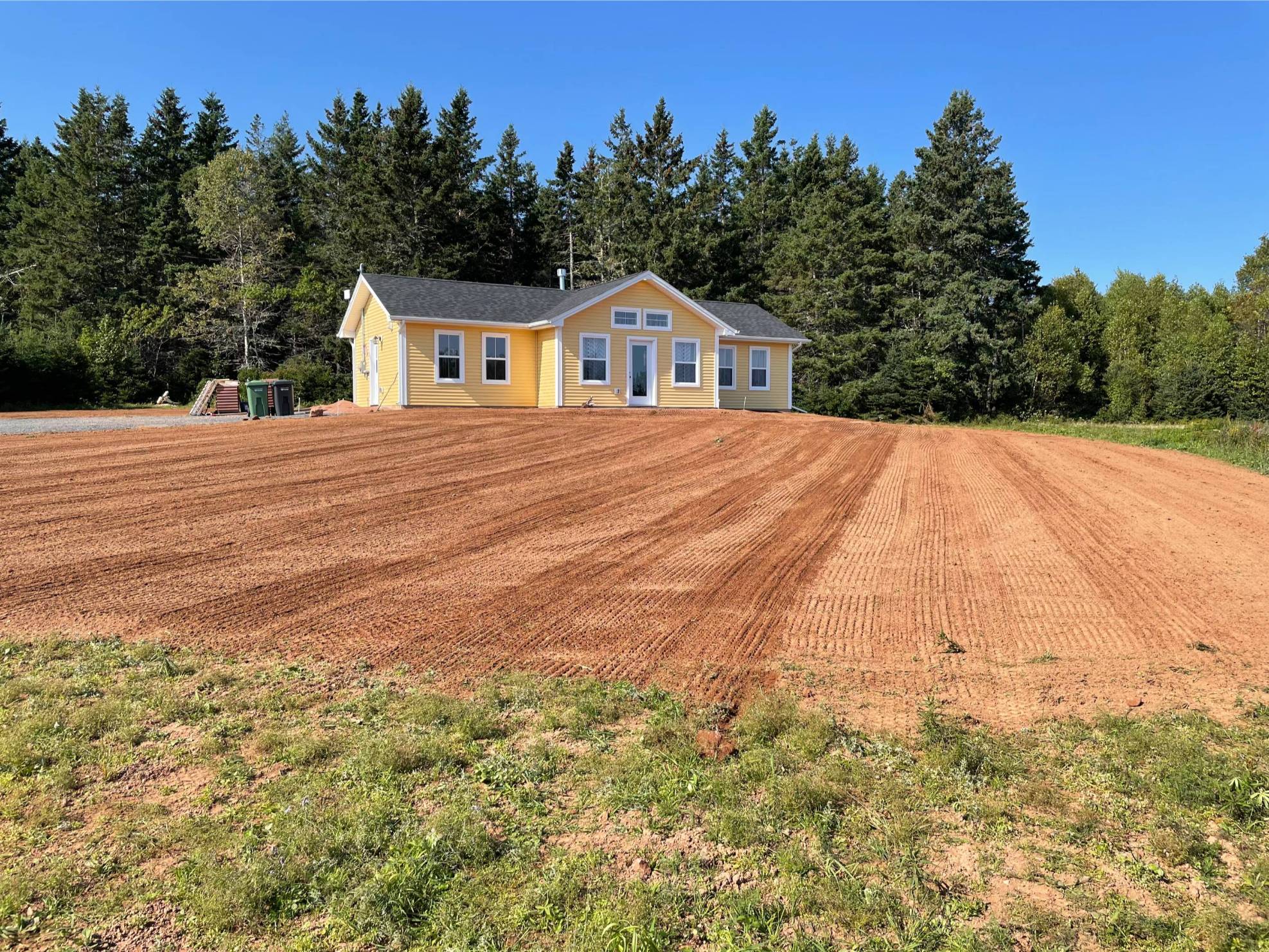 Yard preparation area with freshly graded soil, yellow house in background, surrounded by trees, ready for landscaping and lawn establishment.