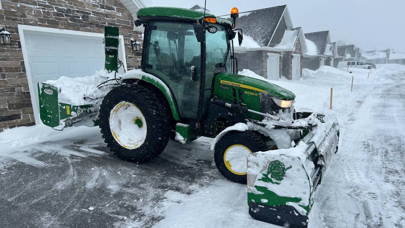 John Deere tractor with snow plow clearing residential driveway covered in snow, emphasizing winter snow removal services by McIsaac Lawncare.