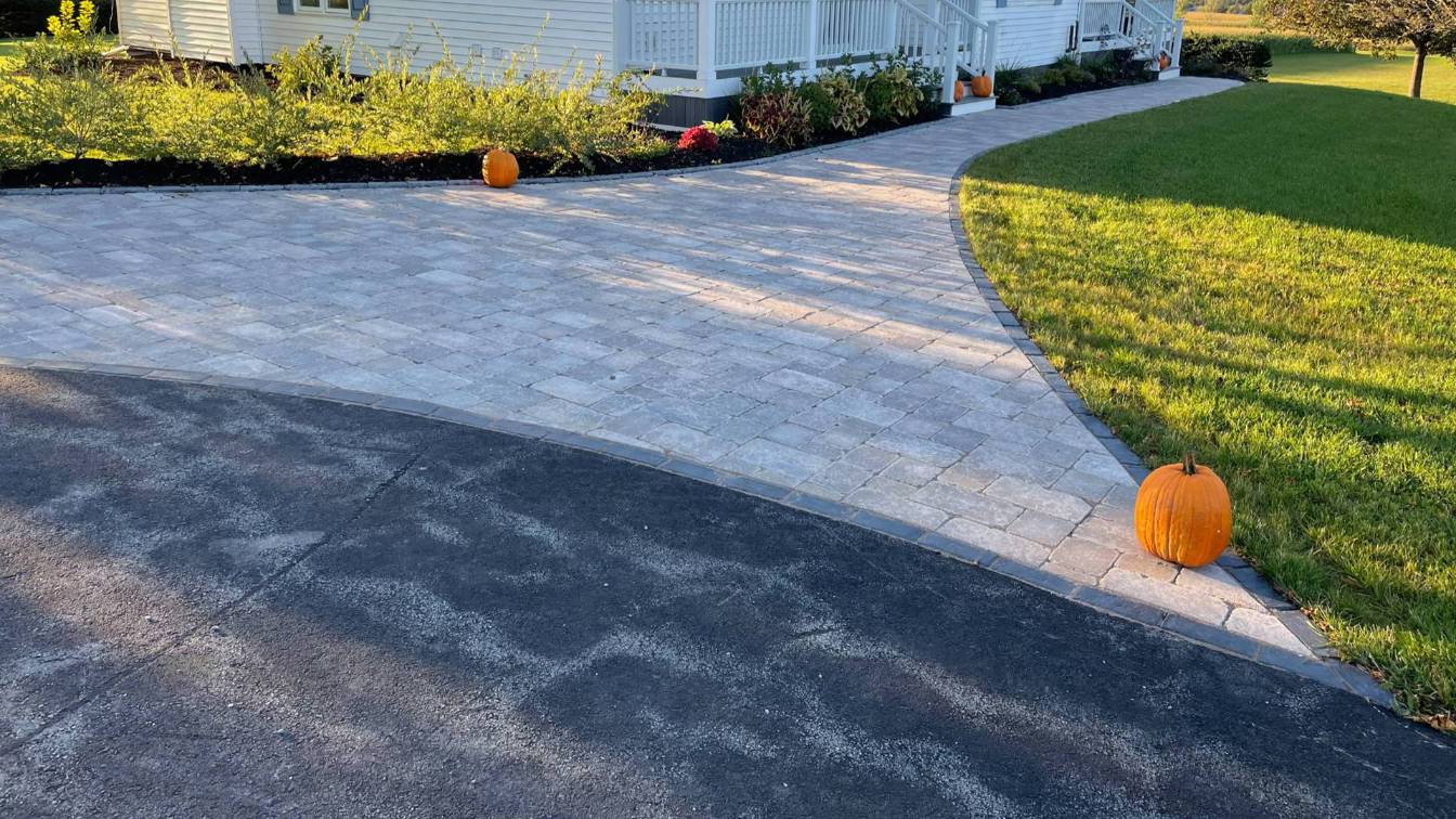 Paver driveway with a curved design, bordered by greenery and decorative pumpkins, enhancing outdoor aesthetics and functionality.