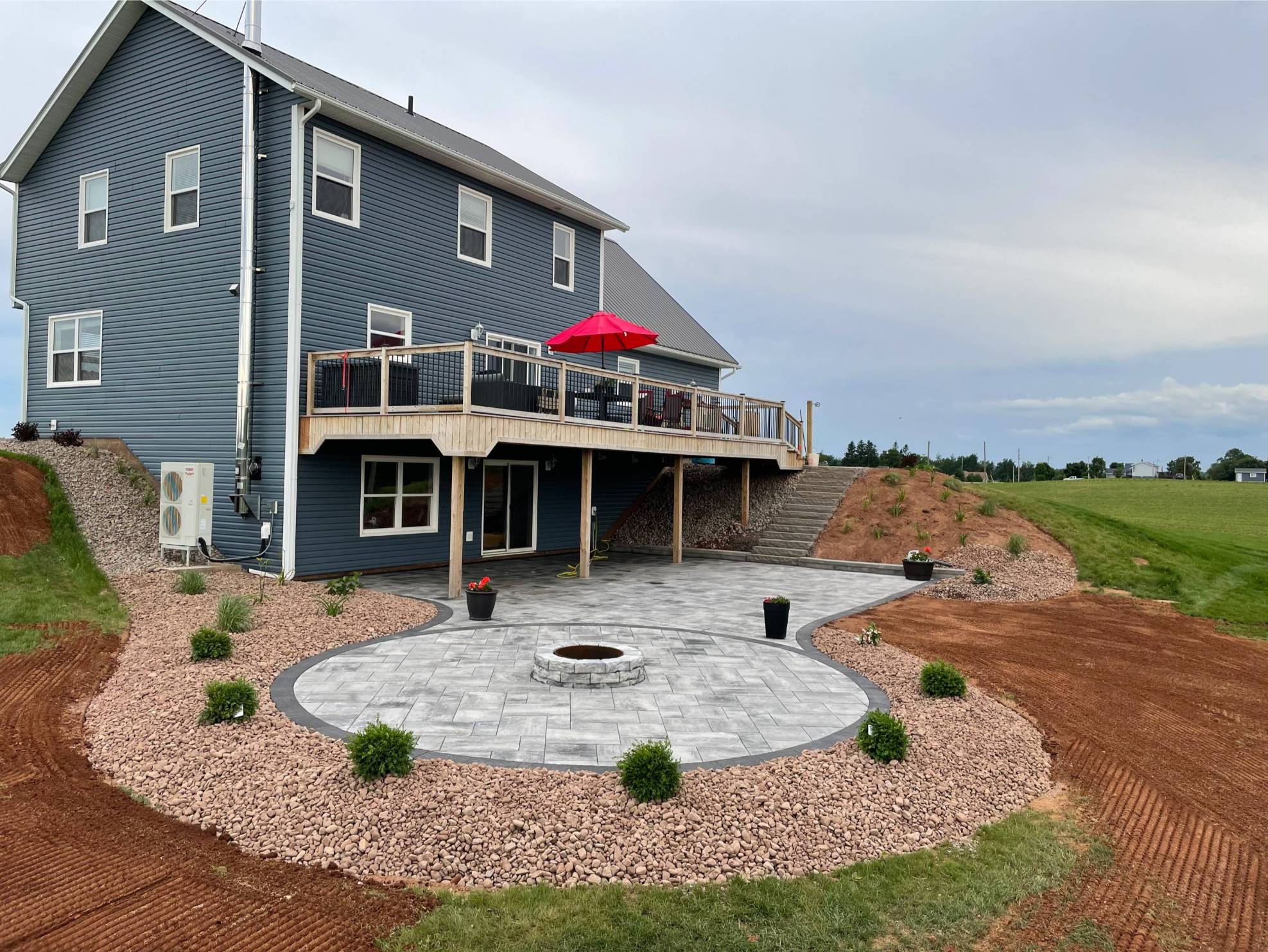 Backyard patio featuring a circular stone fire pit, surrounded by decorative stone landscaping and greenery, adjacent to a modern home with a wooden deck and red umbrella, showcasing functional outdoor living space by McIsaac Lawncare.