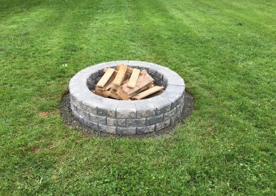 Fire pit with stacked firewood surrounded by a stone circular border in a well-maintained grassy backyard.